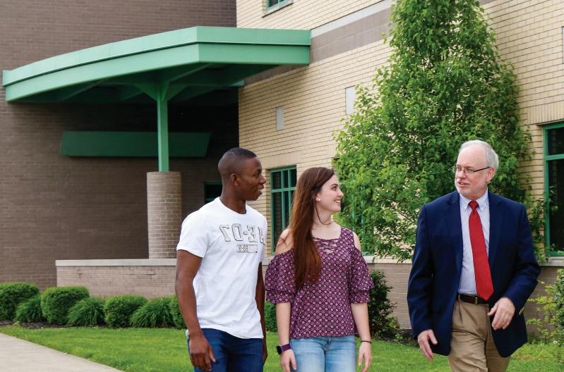 Three people are walking and conversing outside a building with green trim. A man in a suit and tie is accompanied by two young adults, one in a patterned blouse and jeans, and the other in a white T-shirt.